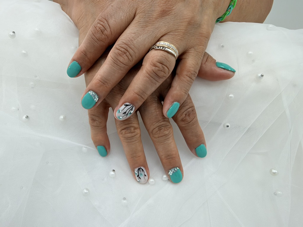 Manicura de Miora Nail: uñas cortas turquesas, flores pintadas sobre blanco y pequeños cristales, con ambas manos visibles